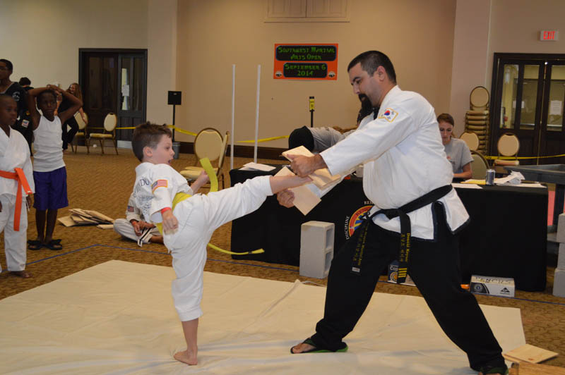 young boy breaks board held by a coach with a kick at martial arts tournament