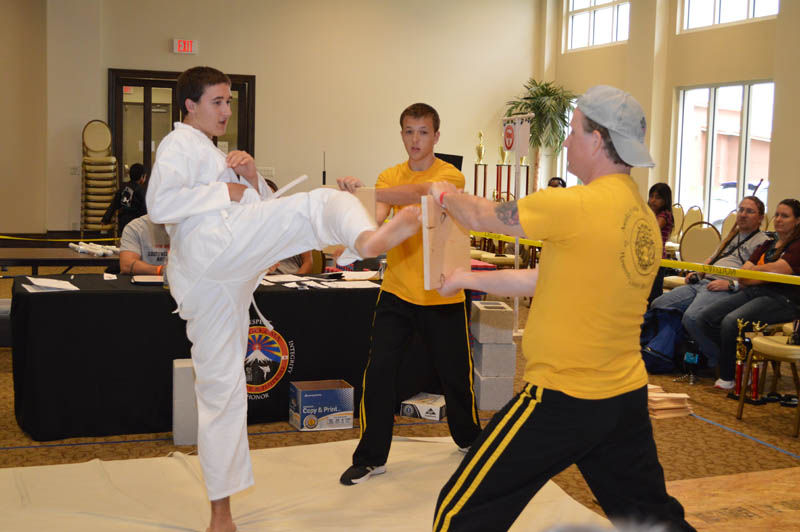 teen boy kicks board held by a coach at martial arts tournament